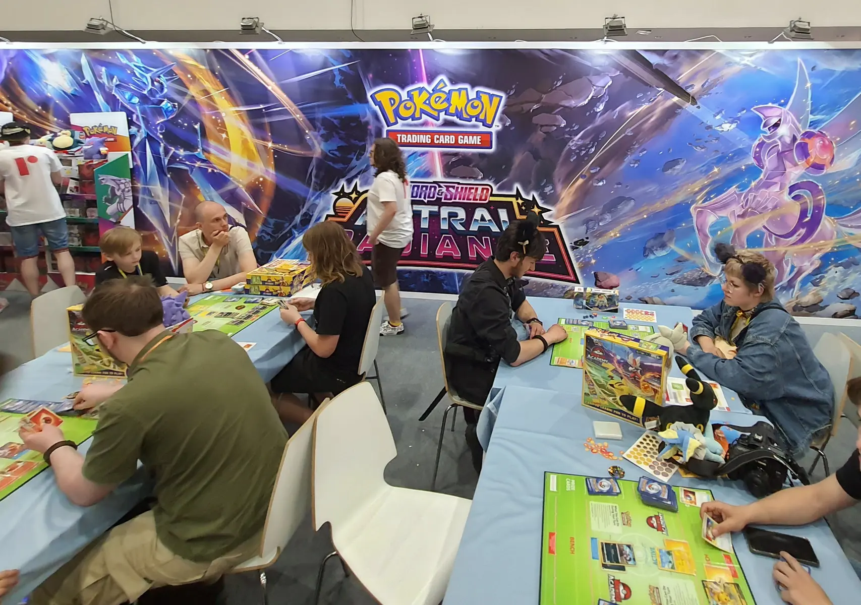 Mensen die het Pokémon Trading Card Game spelen tijdens een evenement voor een grote Pokémon-achtergrond.